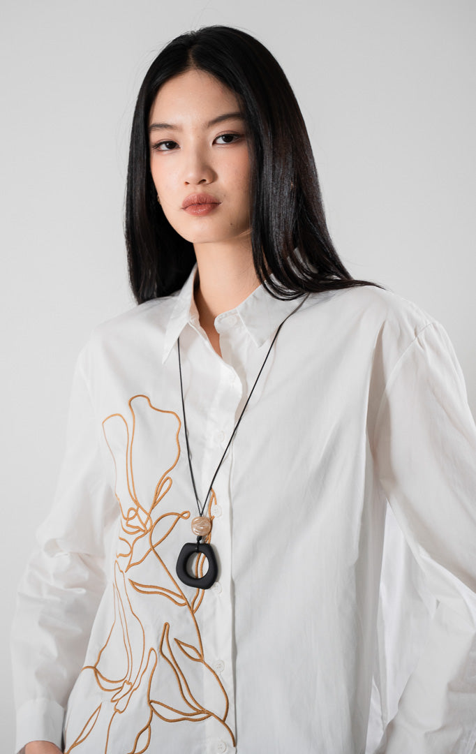 kemeja lengan panjang berwarna putih Adela Top White berbahan katun dengan detail bordir bunga estetis di bagian depan, dipadukan dengan celana cokelat.