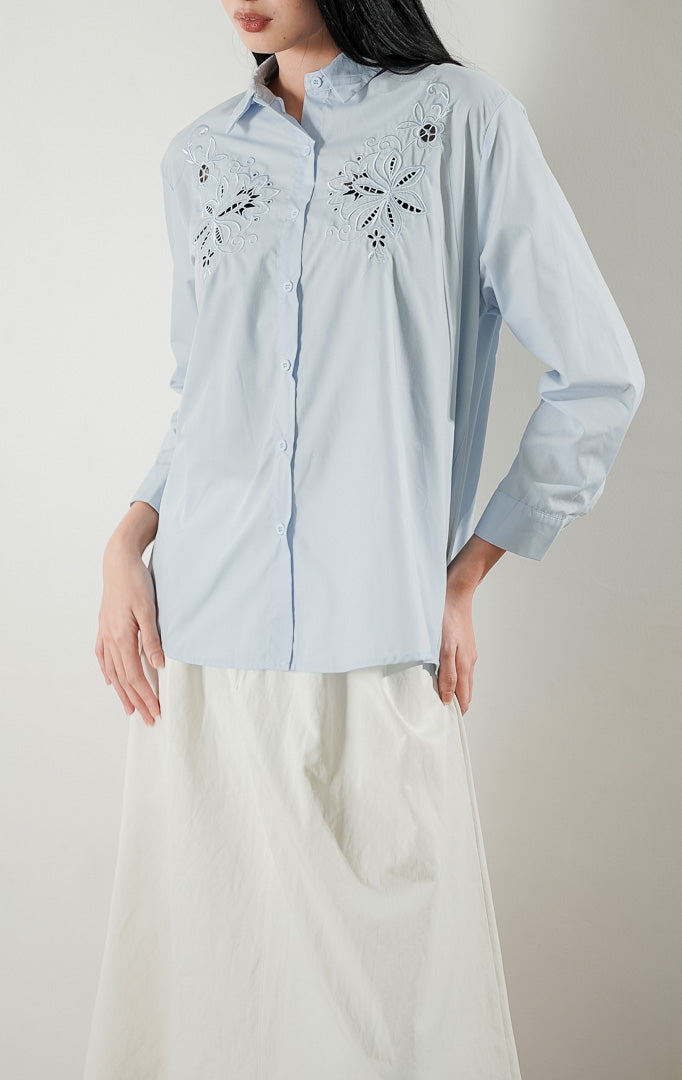 kemeja lengan panjang berwarna biru muda Amaranth Top Blue berbahan katun dengan detail bordir bunga di bagian dada, dipadukan dengan rok panjang berwarna putih.