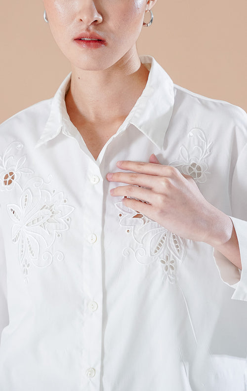 kemeja lengan panjang berwarna putih Amaranth Top White berbahan katun dengan detail bordir bunga di dada, dipadukan dengan rok denim panjang.