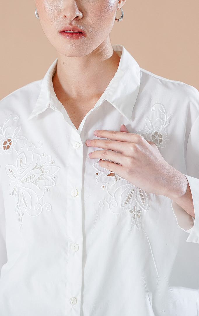 kemeja lengan panjang berwarna putih Amaranth Top White berbahan katun dengan detail bordir bunga di dada, dipadukan dengan rok denim panjang.