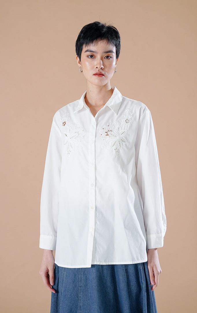 kemeja lengan panjang berwarna putih Amaranth Top White berbahan katun dengan detail bordir bunga di dada, dipadukan dengan rok denim panjang.