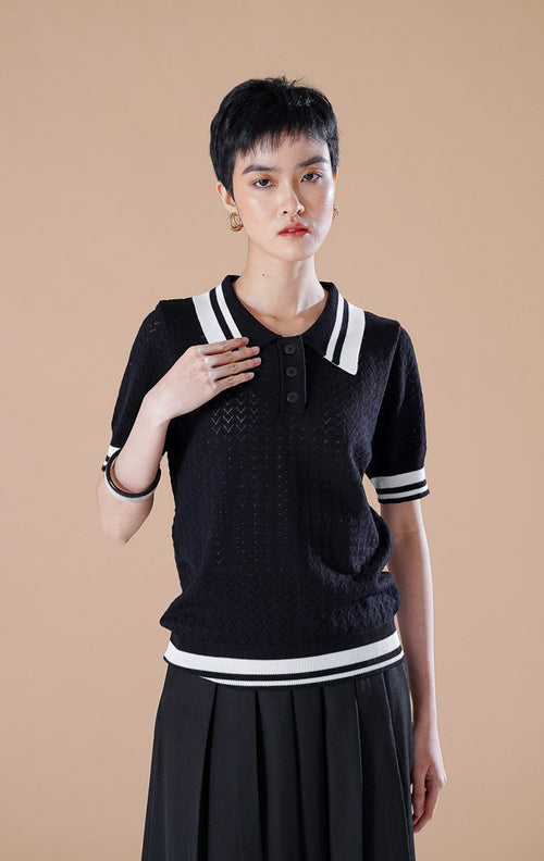 Amie Top Black, atasan polo rajut lengan pendek warna hitam dengan detail tekstur pointelle dan garis aksen putih pada kerah serta ujung lengan, dipadukan rok lipit hitam.