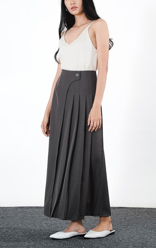 Azalia Skirt warna dark grey, rok panjang plisket berbahan poliester dengan aksen kancing asimetris di pinggang, dipadukan dengan sepatu putih.