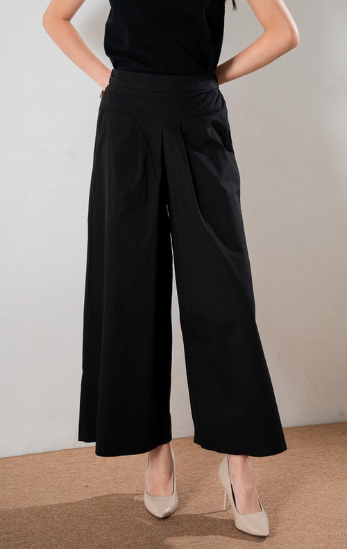 Barbara Pants Black, celana panjang wide leg hitam dengan detail lipit (pleats) depan yang rapi, dipadukan dengan atasan tanpa lengan hitam dan sepatu hak tinggi krem.