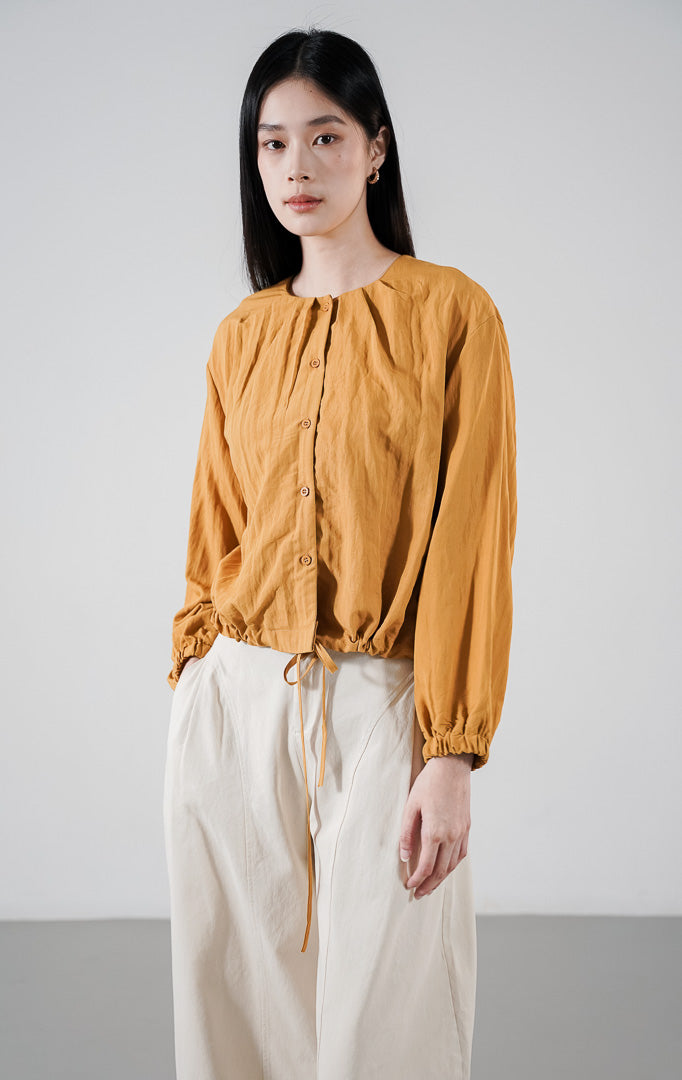 Callum Top warna mustard, atasan lengan panjang berkerah bulat dengan kancing depan dan detail tali serut (drawstring) di pinggang, dipadukan dengan celana putih tulang.