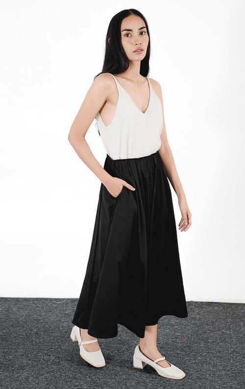 Catarina Skirt warna hitam, rok midi potongan A-line dengan detail lipit (gathered) dan saku samping, dipadukan dengan sepatu Mary Jane putih.