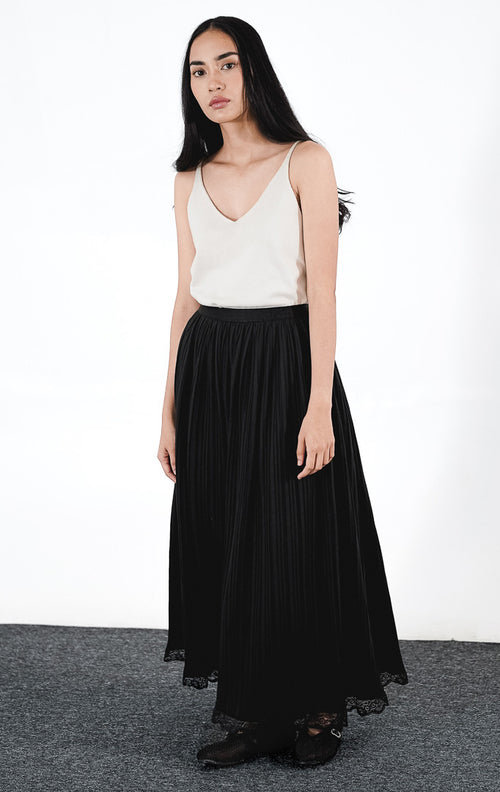 Cressida Skirt warna hitam, rok midi plisket (pleated) berpotongan rapi dengan detail ujung renda di bagian bawah, dipadukan dengan tank top putih.
