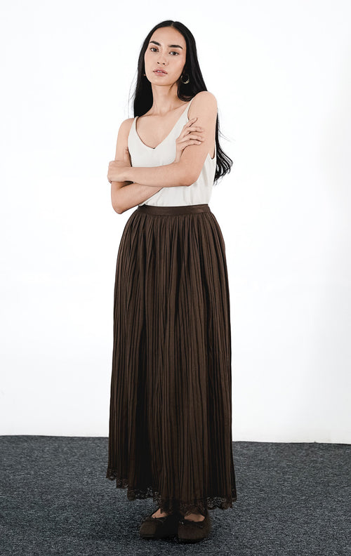 Cressida Skirt warna cokelat (brown), rok midi plisket (pleated) berpotongan rapi dengan detail ujung renda di bagian bawah, dipadukan dengan atasan tanpa lengan warna terang.
