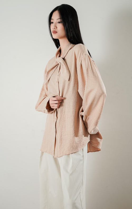 kemeja lengan panjang berwarna khaki atau krem Cristina Top berbahan katun dengan detail kerah pita di bagian leher, dipadukan dengan celana kulot cream dan sepatu heels silver