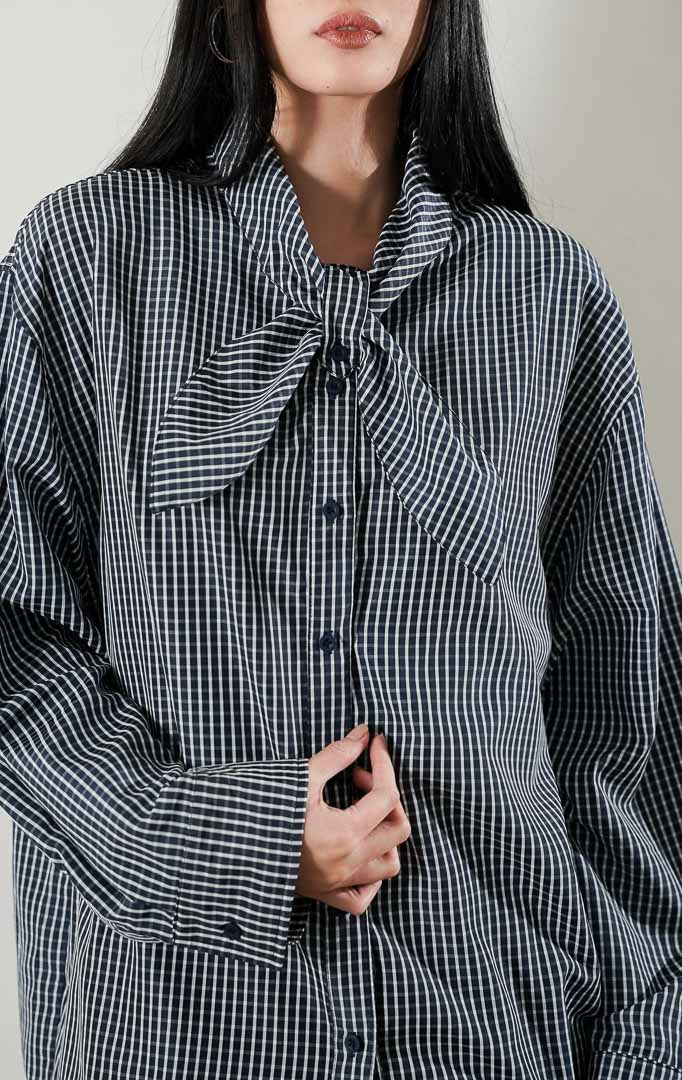Cristina Top Plaid Navy, kemeja katun lengan panjang motif kotak kotak biru dongker dengan detail kerah pita, dipadukan dengan rok midi warna krem.