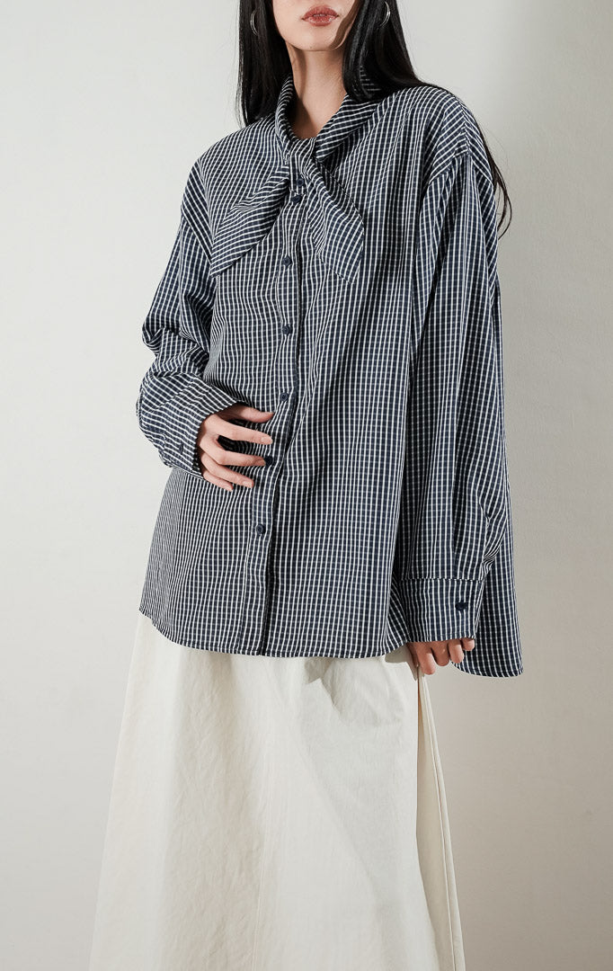 Cristina Top Plaid Navy, kemeja katun lengan panjang motif kotak kotak biru dongker dengan detail kerah pita, dipadukan dengan rok midi warna krem.