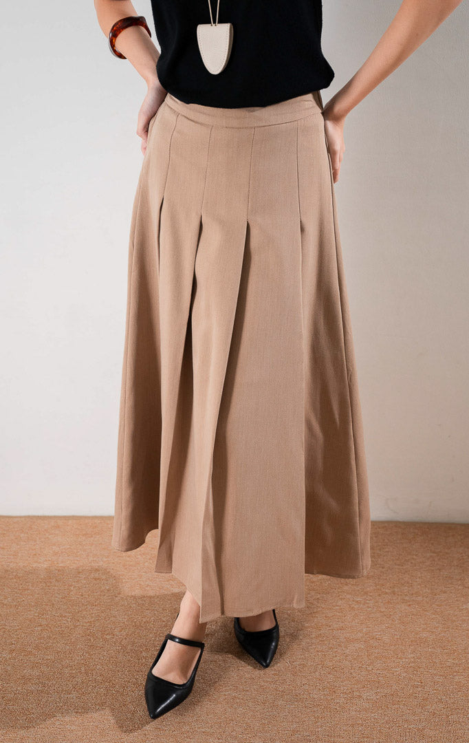 Darana Skirt Brown, rok panjang A line warna cokelat dengan detail lipit (pleats) depan yang rapi, dipadukan dengan tank top hitam dan sepatu hak tinggi hitam.