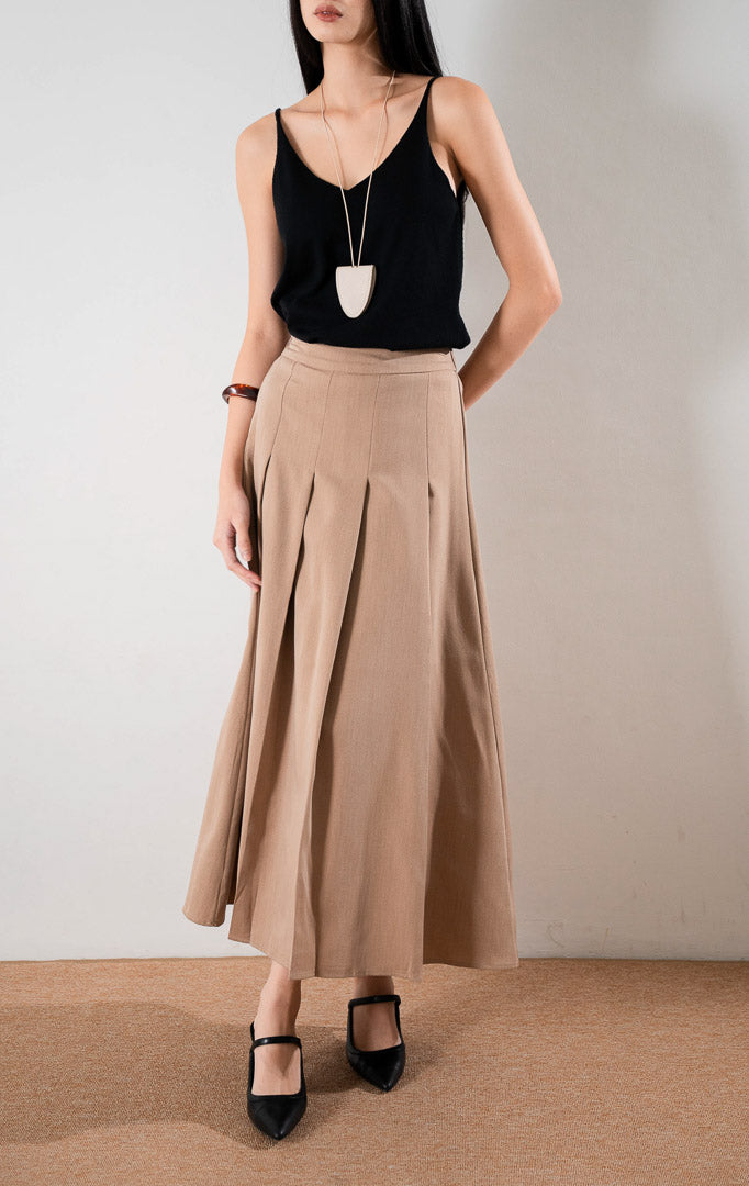 Darana Skirt Brown, rok panjang A line warna cokelat dengan detail lipit (pleats) depan yang rapi, dipadukan dengan tank top hitam dan sepatu hak tinggi hitam.