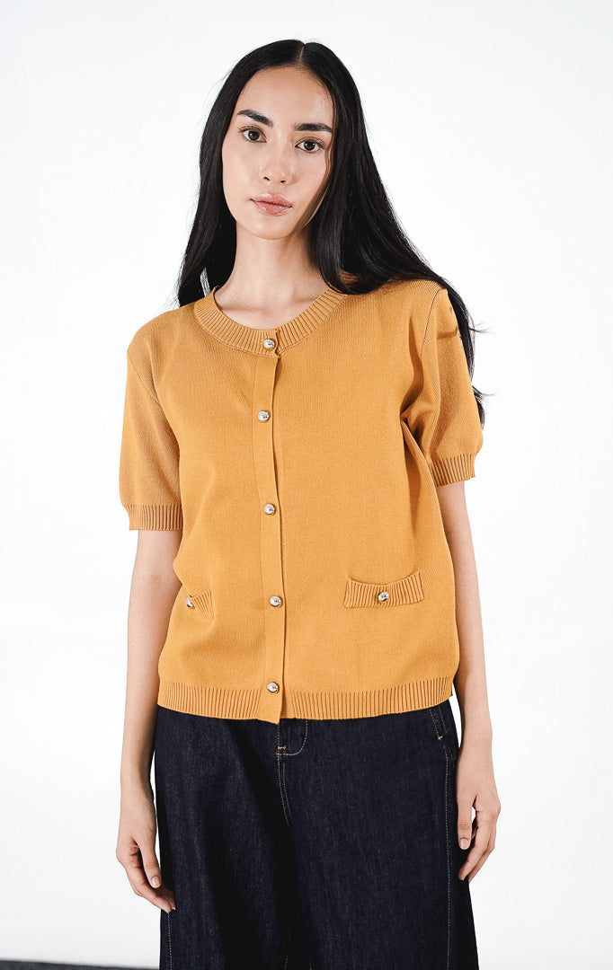 Eliea Top warna mustard, kardigan rajut lengan pendek berkerah bulat dengan detail kancing perak dan dua saku dekoratif di depan.