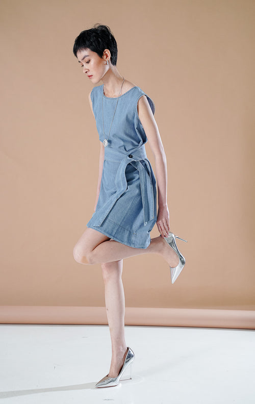Geneva Top Denim, atasan berpotongan tunik panjang atau dress mini tanpa lengan berbahan denim warna biru muda dengan detail sabuk pita di pinggang, dipadukan sepatu hak perak.