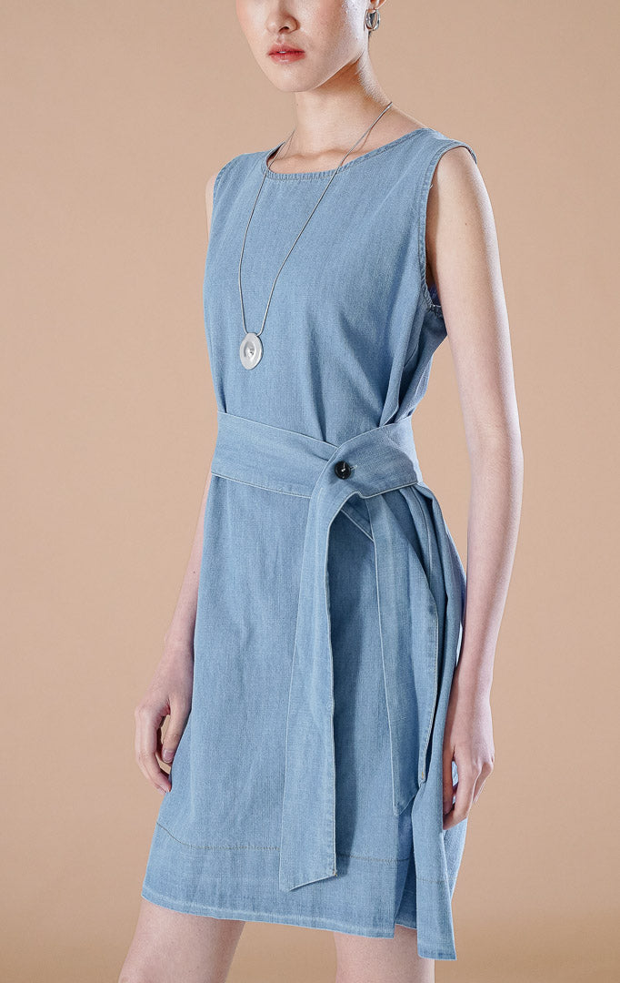 Geneva Top Denim, atasan berpotongan tunik panjang atau dress mini tanpa lengan berbahan denim warna biru muda dengan detail sabuk pita di pinggang, dipadukan sepatu hak perak.