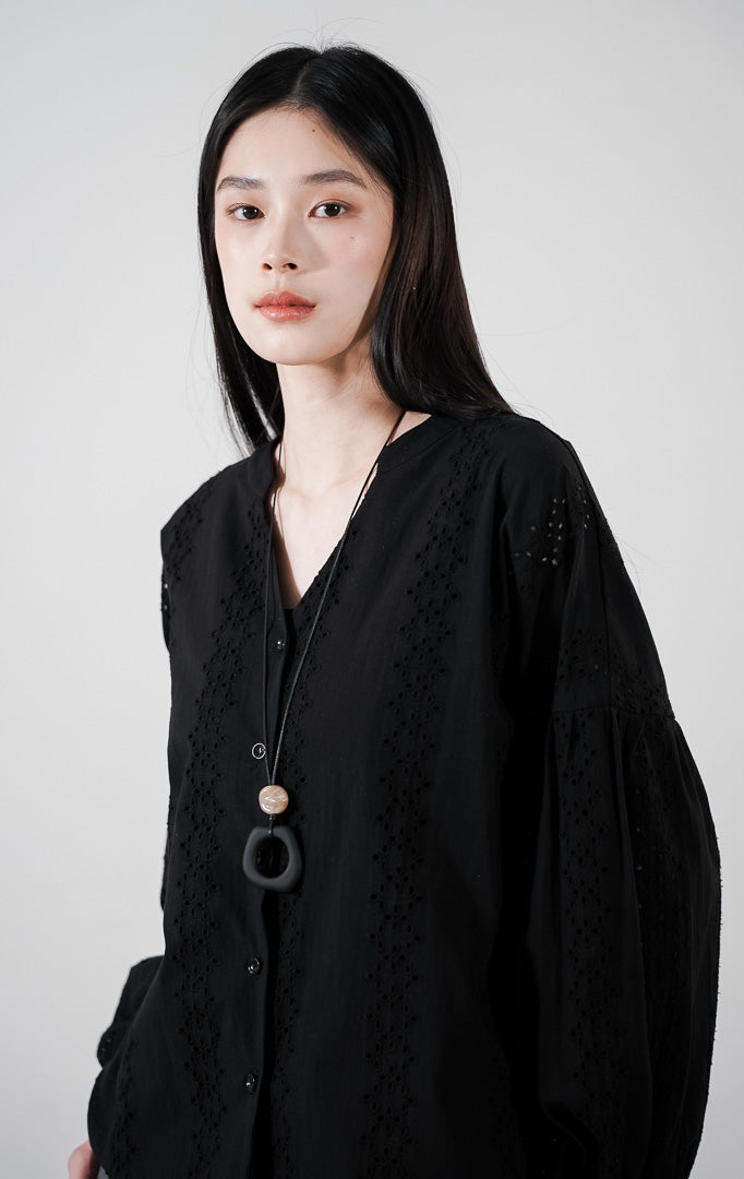 Marcelo Top warna black, atasan katun lengan balon dengan detail bordir eyelet vertikal dan kerah V-neck, dipadukan dengan celana abu-abu.