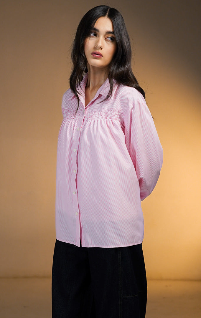 Model wanita mengenakan Osiana Smocked Long Sleeve Shirt warna pink, kemeja lengan panjang motif garis vertikal halus (pinstripe) dengan detail kerutan smock di bagian dada, dipadukan dengan celana denim gelap