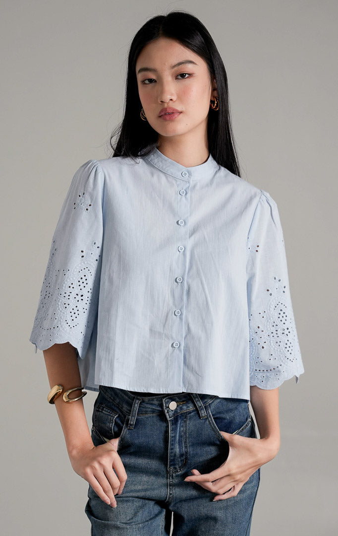 Rochelle Top Light Blue, kemeja katun biru muda dengan detail bordir eyelet pada lengan pendek yang melebar, dipadukan dengan celana jeans wide-leg.