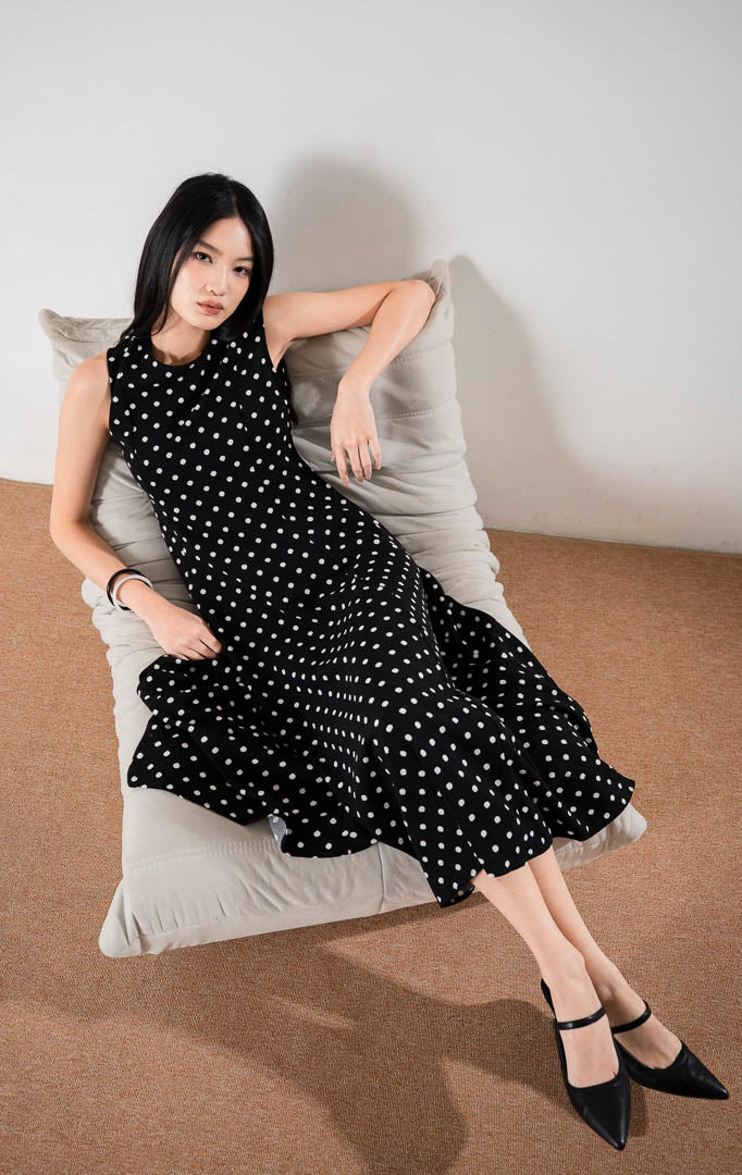 Vianna Dress Black, gaun panjang tanpa lengan motif polka dot putih di atas dasar hitam dengan potongan hem asimetris yang elegan.
