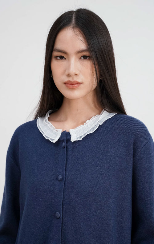 Close-up model mengenakan Aiko Cardigan warna navy, menampilkan detail kerah ruffle lace putih yang kontras dan kancing bungkus rajut di bagian depan.