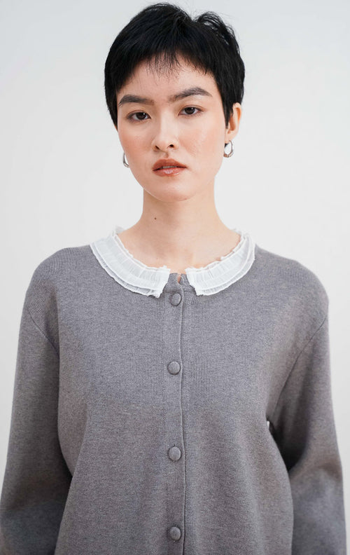 Model mengenakan Aiko Cardigan warna grey, menampilkan detail kerah ruffle lace putih yang cantik, material rajut halus, dan kancing bungkus di bagian depan.