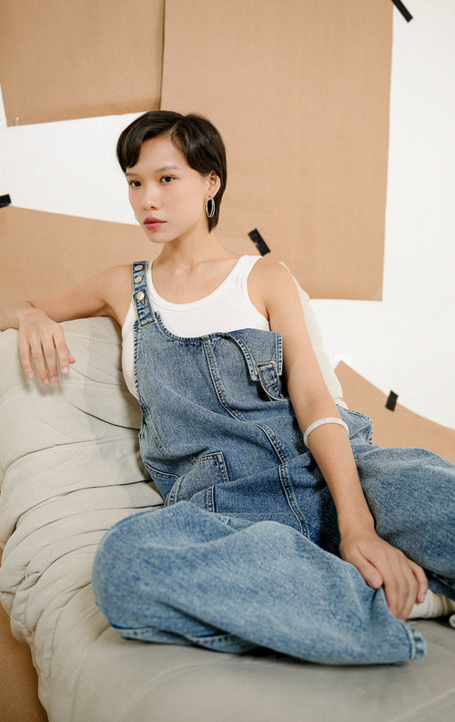 Model berpose duduk mengenakan Amira Pocket Ease Jumpsuit Denim, baju kodok atau jumpsuit jeans wanita dengan potongan santai dipadukan tank top putih.