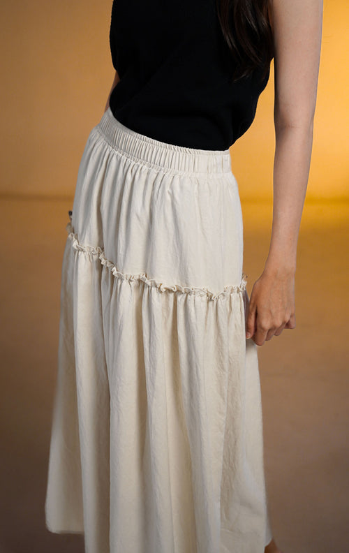 Model wanita mengenakan rok midi susun (tiered skirt) warna ivory bahan peach skin yang flowy dengan atasan tank top hitam.