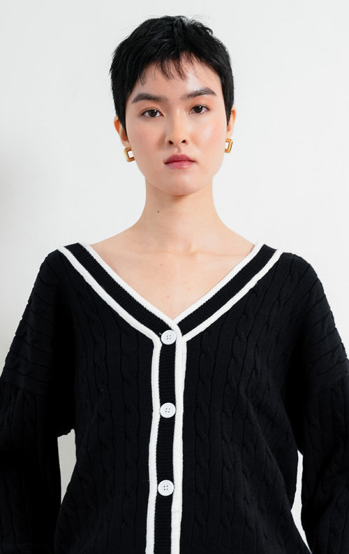 Kardigan rajut wanita lengan panjang warna hitam (black) Annette Cardigan bermotif rajut kepang (cable knit) dengan lis kontras warna putih di leher V (V-neckline) dan bukaan kancing depan.