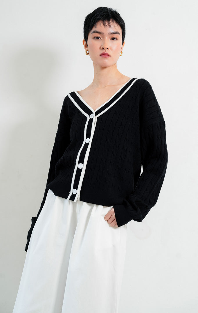 Kardigan rajut wanita lengan panjang warna hitam (black) Annette Cardigan bermotif rajut kepang (cable knit) dengan lis kontras warna putih di leher V (V-neckline) dan bukaan kancing depan.