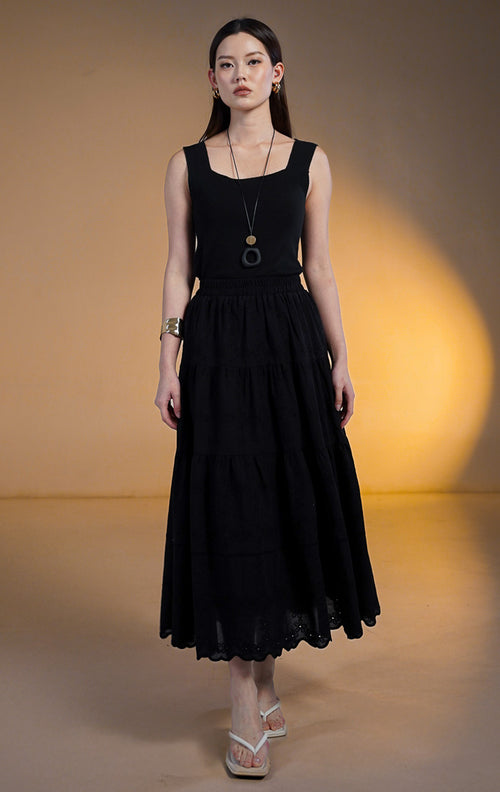 Model wanita mengenakan rok midi hitam model susun (tiered skirt) dengan detail bordir eyelet di bagian bawah, dipadukan dengan tank top hitam dan sandal jepit putih.