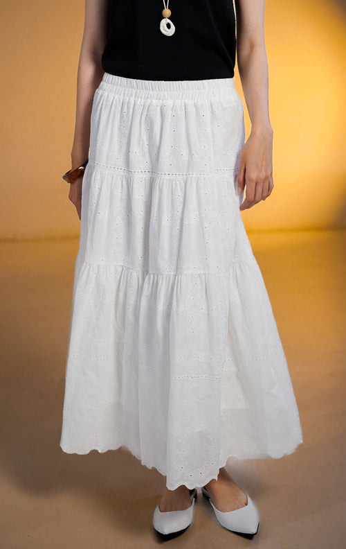 Model wanita mengenakan rok midi putih model susun (tiered skirt) dengan detail bordir eyelet bunga, dipadukan dengan atasan hitam dan sepatu flat putih.