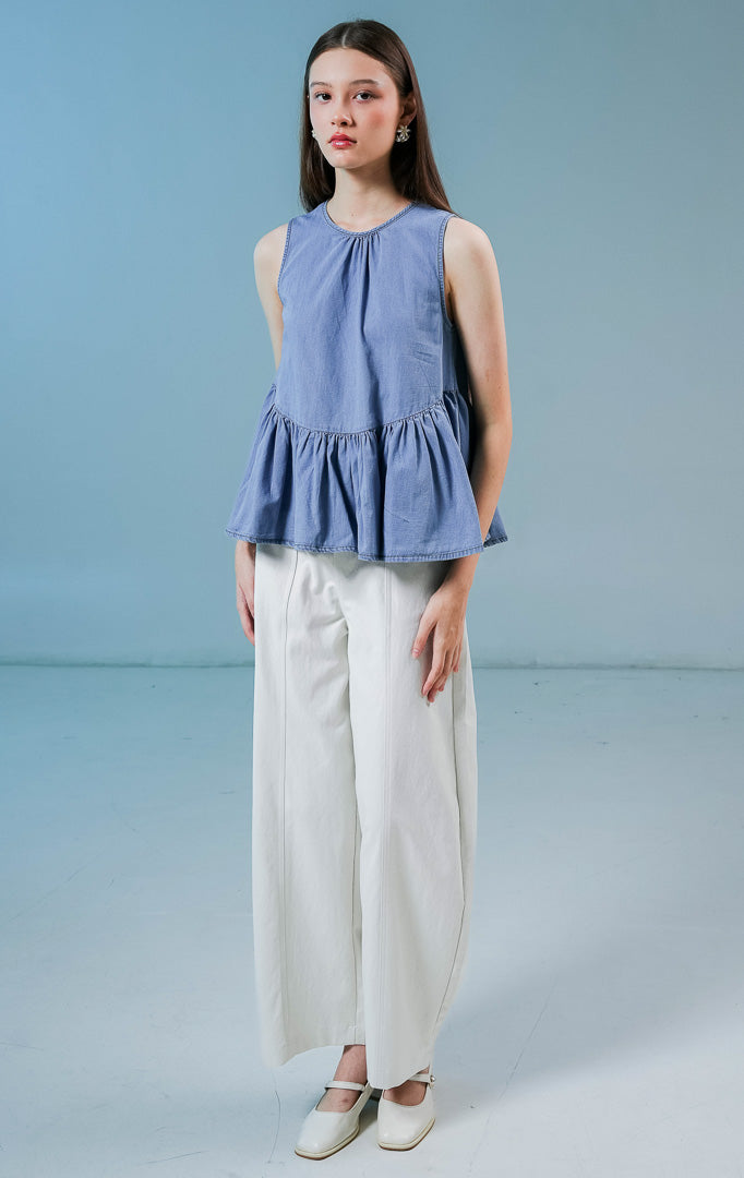Model wanita mengenakan Elvera Sleeveless Peplum Top warna biru denim, atasan tanpa lengan wanita berbahan denim dengan siluet peplum (mengembang di bawah) yang feminin.