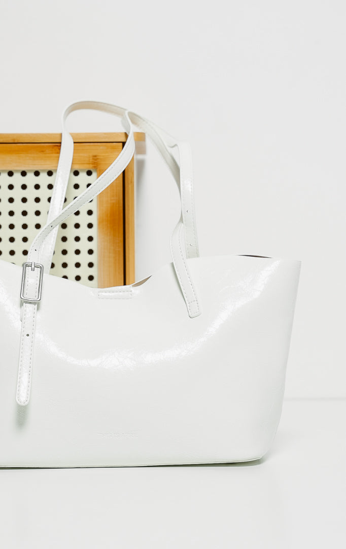 Tas bahu wanita Aure warna putih bersih (white), berdesain siluet trapeze yang melebar ke atas dengan material kulit sintetis mengkilap (glossy finish), menampilkan tali bahu panjang yang dapat diatur menggunakan gesper perak.