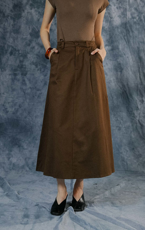 Rok panjang wanita Avella warna dark brown dengan potongan A-line, detail lipit (pleats), kancing depan, dan karet elastis di bagian pinggang belakang.