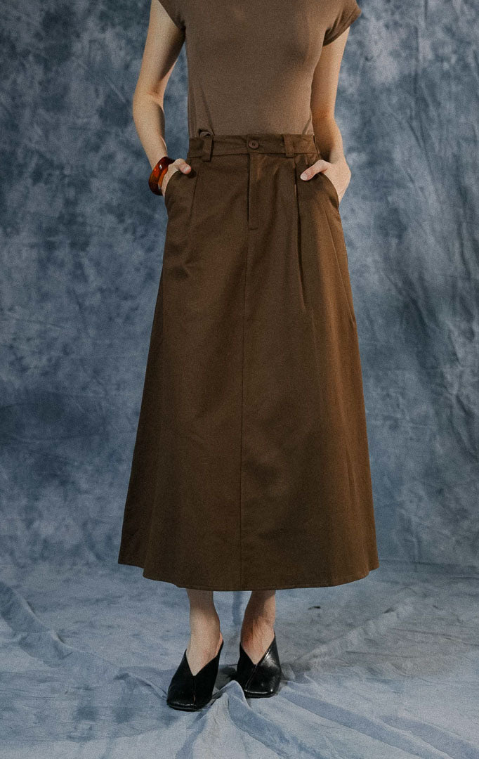 Rok panjang wanita Avella warna dark brown dengan potongan A-line, detail lipit (pleats), kancing depan, dan karet elastis di bagian pinggang belakang.