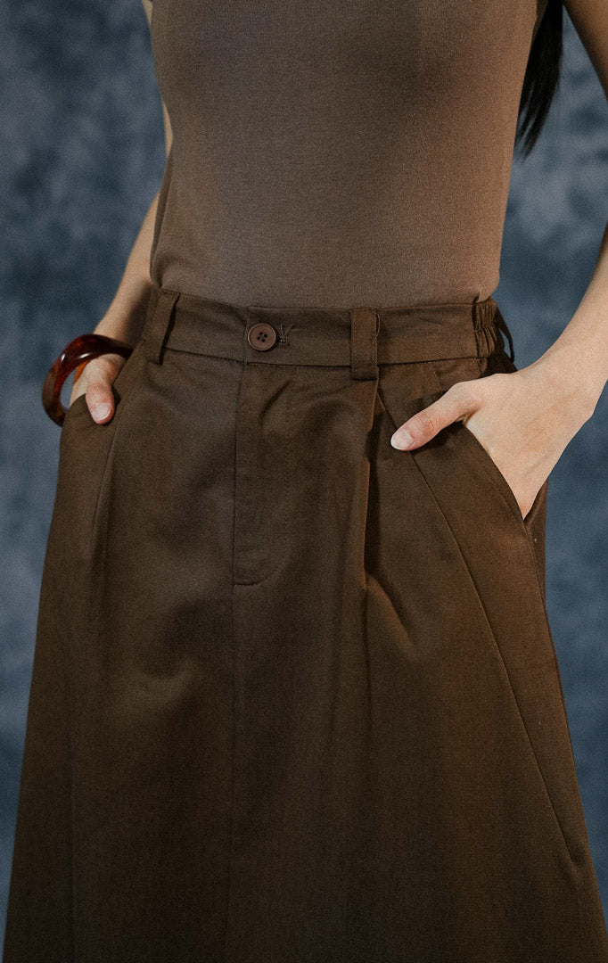 Rok panjang wanita Avella warna dark brown dengan potongan A-line, detail lipit (pleats), kancing depan, dan karet elastis di bagian pinggang belakang.