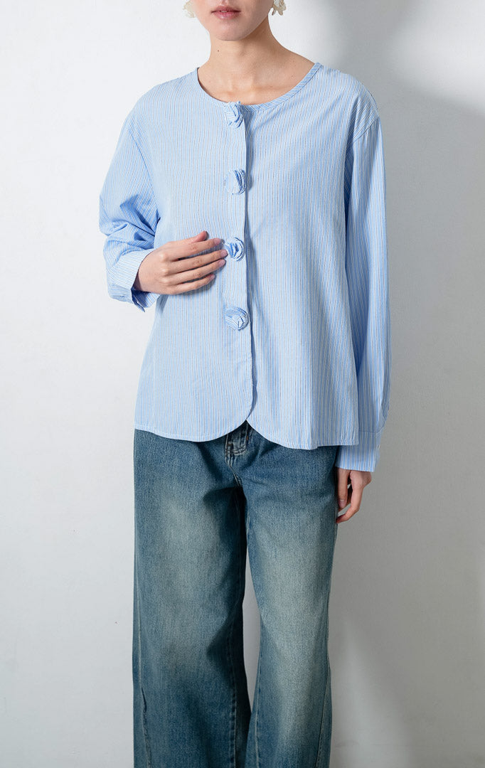 Blus wanita lengan panjang motif garis-garis biru (blue vertical stripes) Azzuria Top dengan kerah bulat (round neckline), detail kancing bungkus berbentuk bunga (floral-shaped fabric-covered buttons), dan kelim melengkung (curved hem).