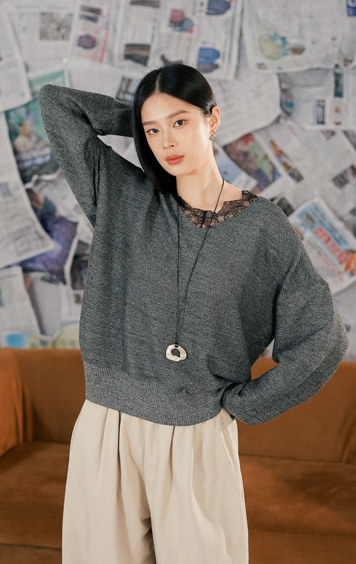 Model wanita mengenakan Bijou Sweater Dark Grey, sebuah atasan rajut lengan panjang warna abu-abu tua. Sweater memiliki potongan leher V-neck dengan detail renda (lace) hitam yang feminin di bagian dada. Dipadukan dengan celana kulot beige longgar untuk tampilan kasual yang nyaman.