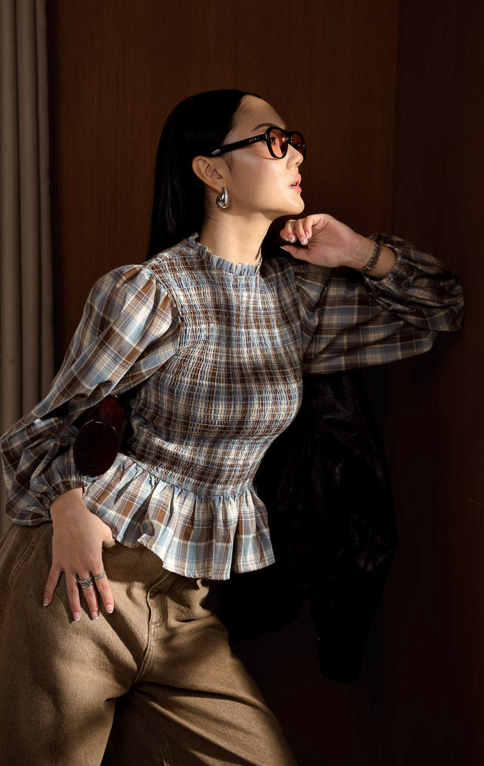 Blouse wanita lengan panjang motif kotak-kotak (plaid blouse) warna cokelat (brown) Bellatrix Top dengan garis leher mock neck beraksen rumbai halus (refined mock neckline with delicate frill detailing), detail smock di bagian depan, serta bahan katun poliester berstruktur.