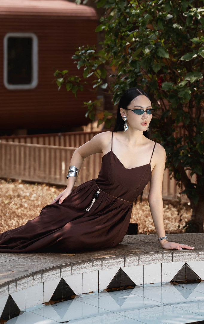 Dress panjang wanita warna cokelat tua (dark brown camisole maxi dress) Daphne Dress dengan model V-neck, tali bahu dapat disesuaikan (adjustable straps), serta bahan campuran katun poliester berstruktur dengan kerut elastis di bagian belakang.