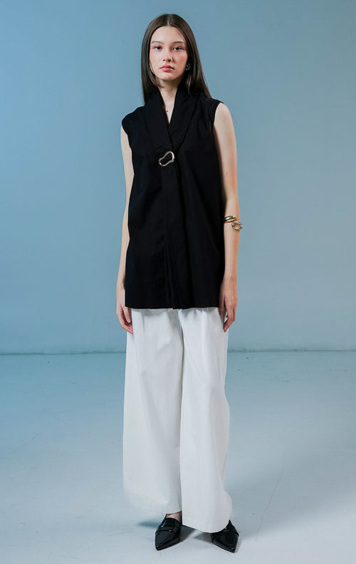 Model wanita mengenakan Fayola Shawl Collar Top warna hitam, atasan tanpa lengan berbahan katun dengan siluet drape yang elegan dan aksen gesper metalik di bagian dada.