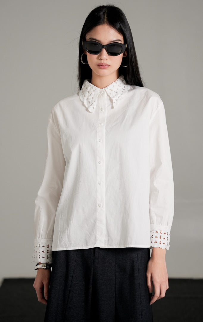 kemeja lengan panjang Brigita Top berwarna putih berbahan katun dengan detail kerah dan ujung lengan beraksen crochet, dipadukan dengan rok panjang hitam dan kacamata gelap.