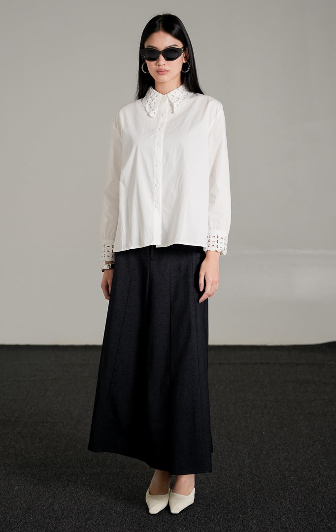 kemeja lengan panjang Brigita Top berwarna putih berbahan katun dengan detail kerah dan ujung lengan beraksen crochet, dipadukan dengan rok panjang hitam dan kacamata gelap.