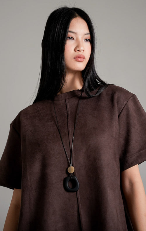 Callidora Suede Top lengan pendek warna cokelat tua bersiluet longgar, dipadukan dengan celana panjang gading, kalung panjang, dan sepatu mules hitam.