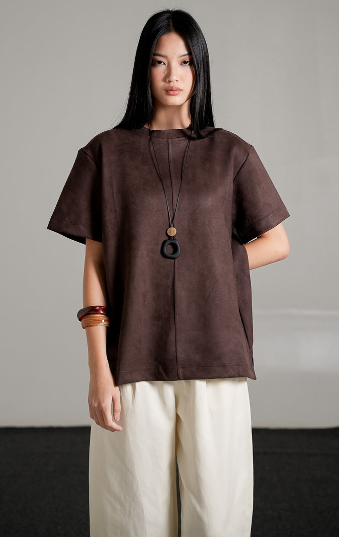 Callidora Suede Top lengan pendek warna cokelat tua bersiluet longgar, dipadukan dengan celana panjang gading, kalung panjang, dan sepatu mules hitam.