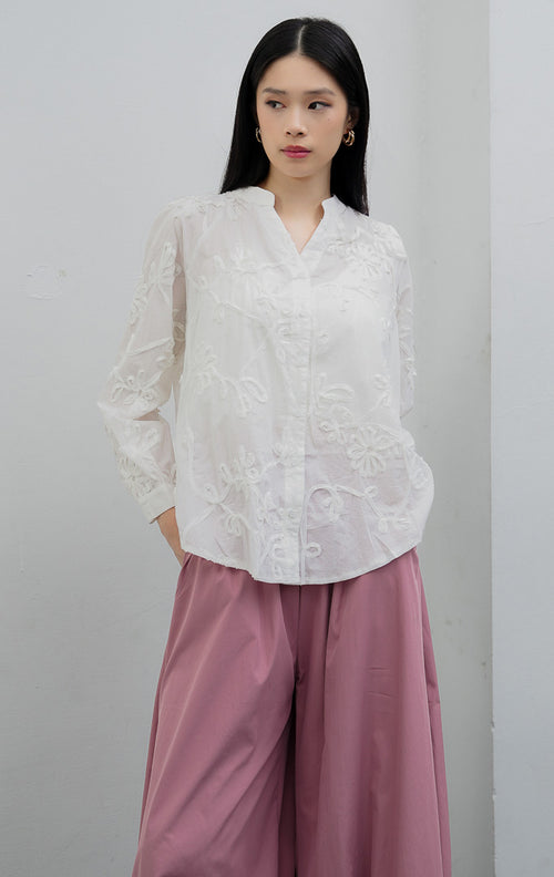 Blus wanita lengan panjang warna putih (white) Camilla Top berbahan katun ringan (lightweight cotton) dengan aksen aplikasi bordir bunga (floral appliqué detailing), efek semi-transparan (subtle sheer), dan bukaan kancing depan.