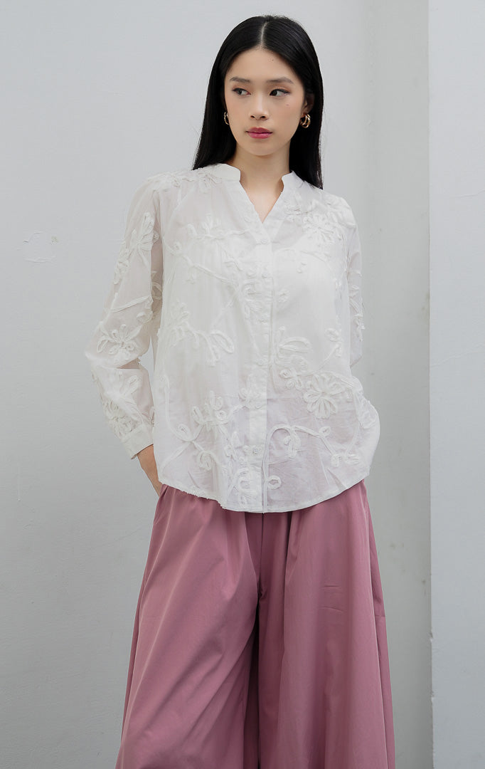 Blus wanita lengan panjang warna putih (white) Camilla Top berbahan katun ringan (lightweight cotton) dengan aksen aplikasi bordir bunga (floral appliqué detailing), efek semi-transparan (subtle sheer), dan bukaan kancing depan.