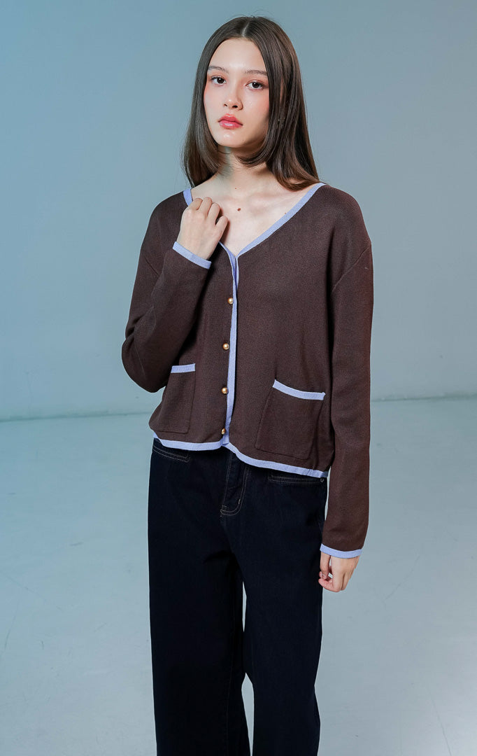 Model wanita mengenakan Darien V-Neck Cardigan warna coklat tua, luaran cardigan rajut wanita dengan detail garis tepi kontras berwarna biru muda, saku depan, dan kancing.
