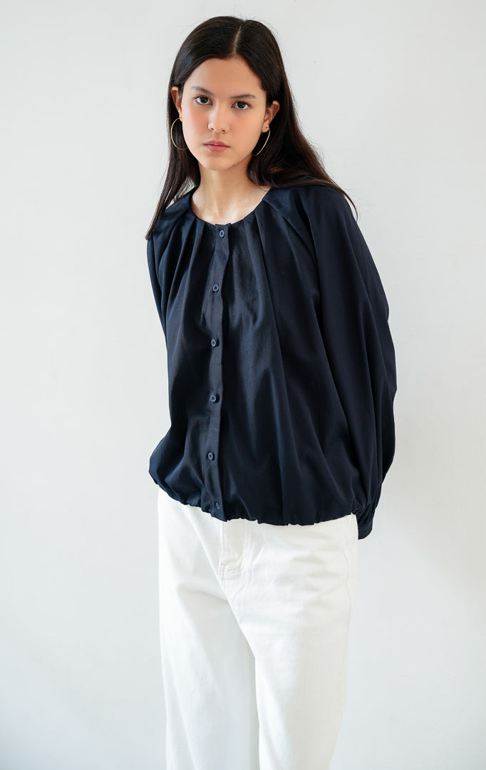 Atasan wanita lengan panjang warna biru navy (navy blue) Catharine Top berbahan katun twill (cotton twill) dengan kerah bulat, bukaan kancing depan, dan detail tali serut di bagian bawah (drawstring hem).